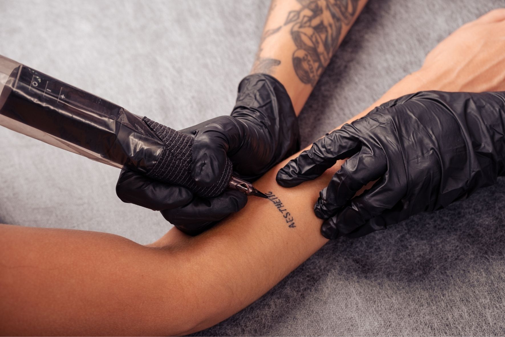 Main gantée utilisant une machine à tatouer sur l’avant-bras d’une personne dans un environnement de tatouage  à Val-David